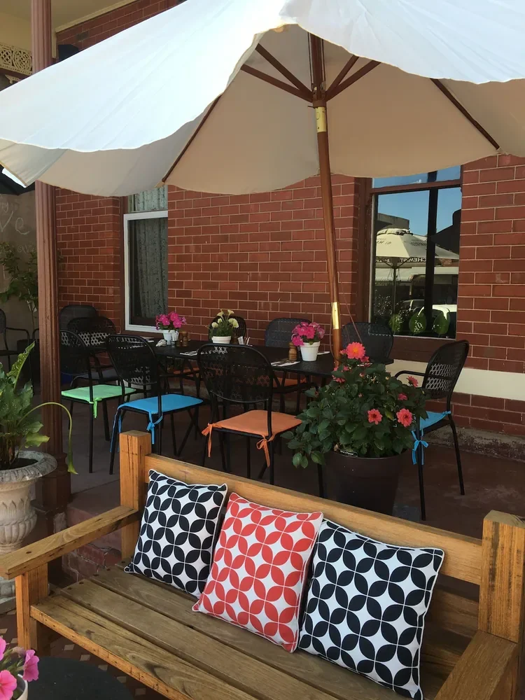 Tables under a white umbrella on the verandah, with a timber bench, patterned cushions and flowers